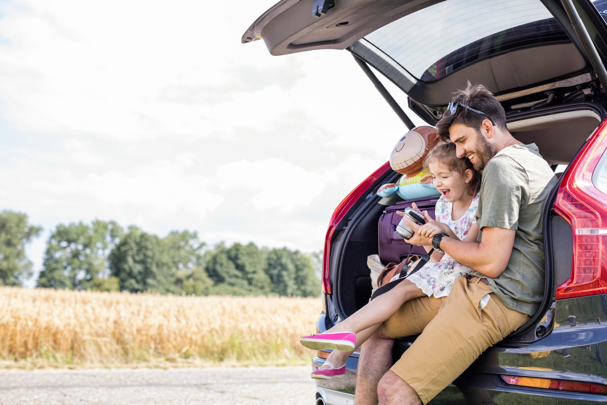 Father and daughter sitting in open car boot looking at camera at break of a road trip