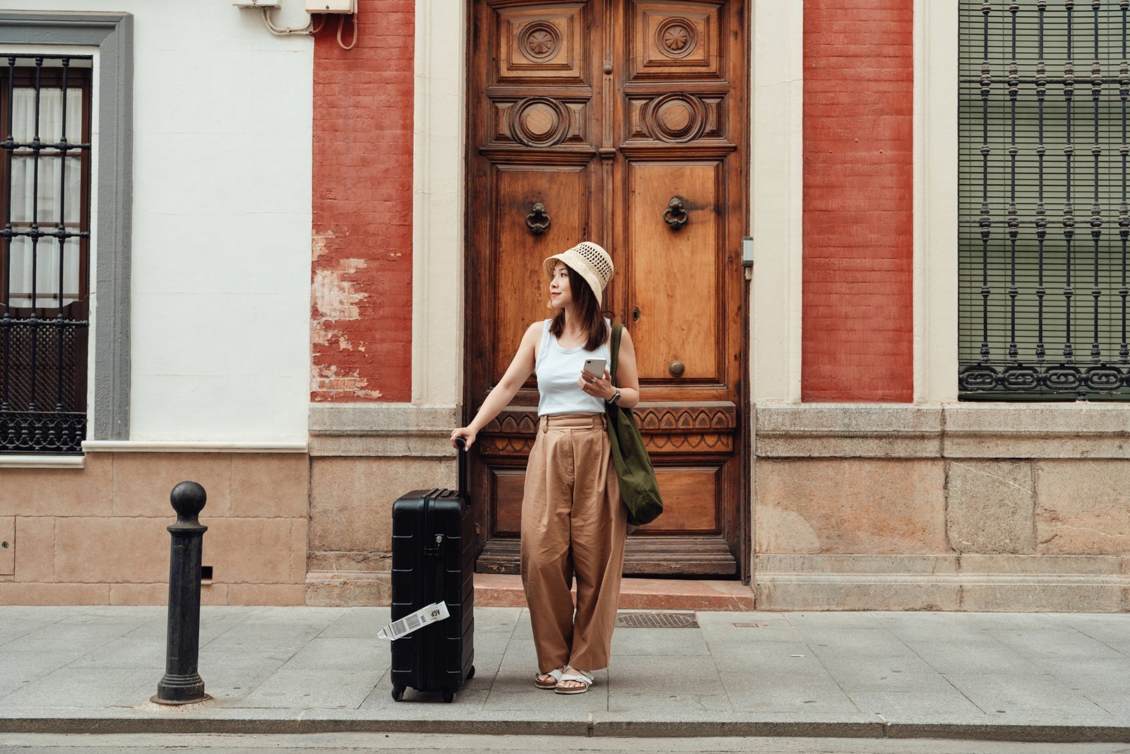 Full length of young Asian woman with luggage ordering taxi pick-up service on smartphone